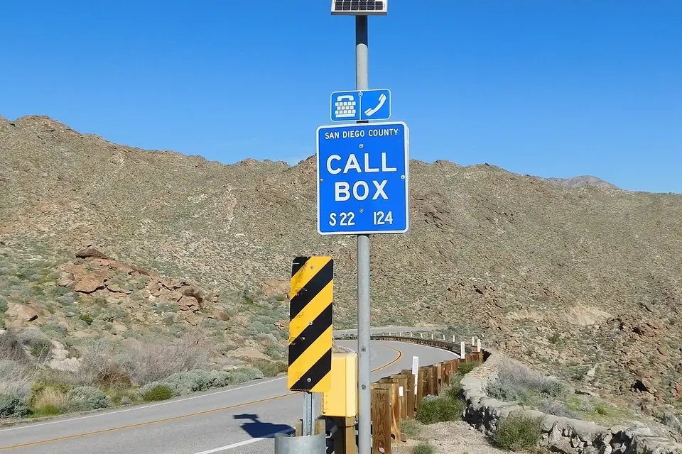 A blue roadside call box sign on a pole with a small solar panel, beside a desert highway