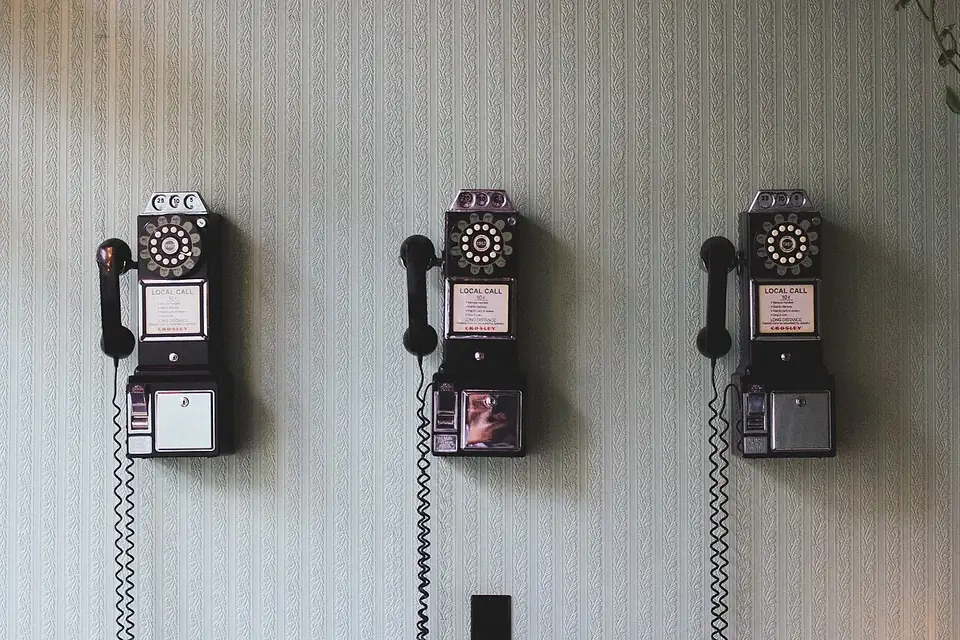 Three vintage pay telephones with rotary dials and coiled cords mounted in a row on a pale wall