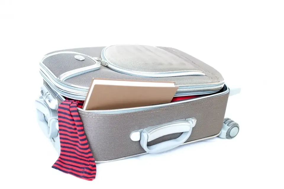 An open grey suitcase on wheels, half packed, with a book and a striped scarf spilling out