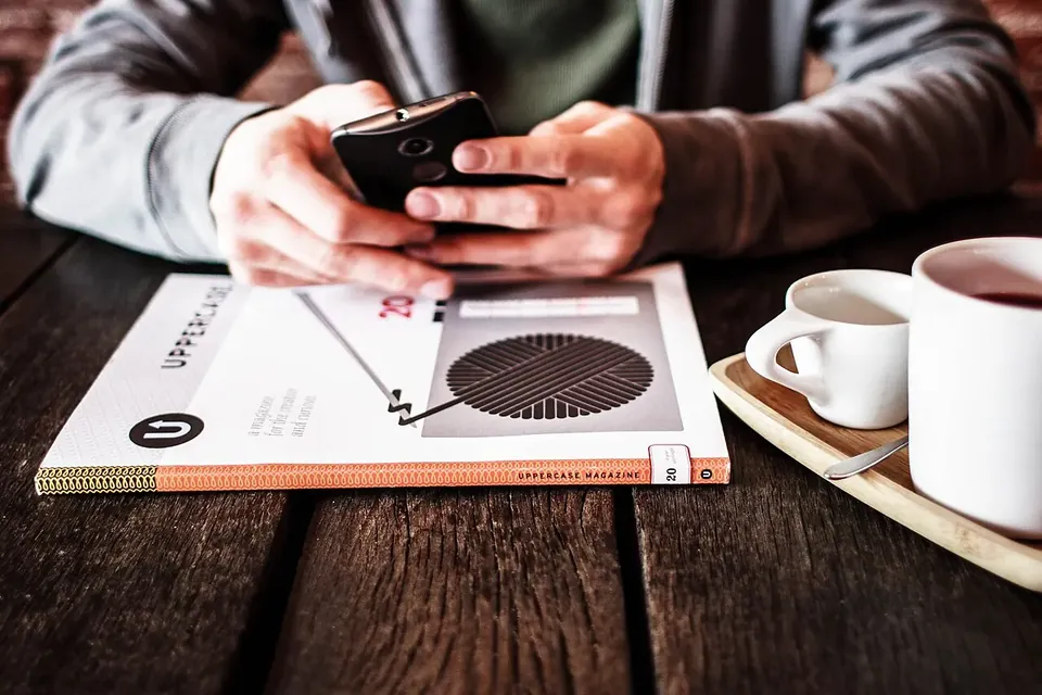 Two hands holding a smartphone over a café table with a magazine and two cups of coffee
